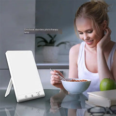 Woman sitting at a table with a bowl of cereal, next to a light therapy lamp, in a kitchen setting.